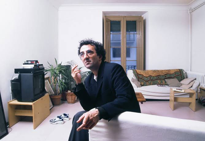 Roberto Bolaño sitting on the arm of a sofa in his flat in Blanes, cigarette in hand, a television and a plant behind him
